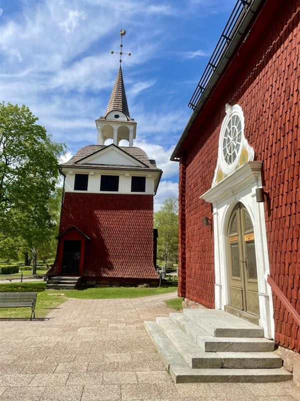 Sundborns kyrka, exteriörbild, röd träkyrka med grön dörr, vitt träsnideri runt, klockstapel rakt fram.
