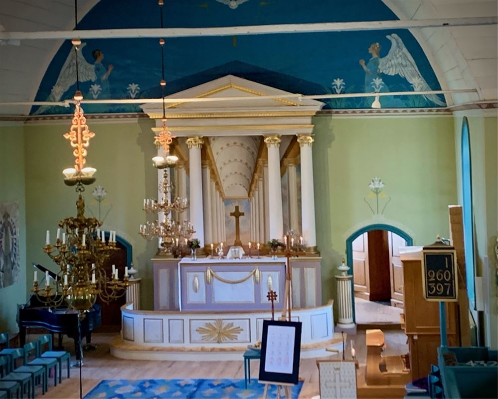 Interiörbild på altaret, ljusa väggar och tak, blå Carl Larssonmålning ovanför altaret.