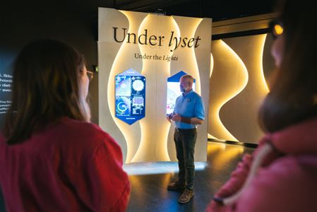 Man in front of an exhibition with illuminated panels and two people listening.