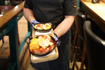 Chef carrying two small cast iron pans on a serving board, with a hot dish and dessert, in a restaurant with bar stools and tables in the background.