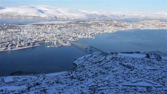 Luftfoto av snødekt Tromsø med bro og fjell i bakgrunnen.